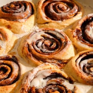 Close-up of freshly baked vegan protein cinnamon rolls topped with a silky vanilla coconut cream icing. The soft, fluffy cinnamon rolls feature thick cinnamon sugar swirls with golden-brown edges, while the warm icing melts into every layer, creating a rich, gooey texture perfect for breakfast or dessert.