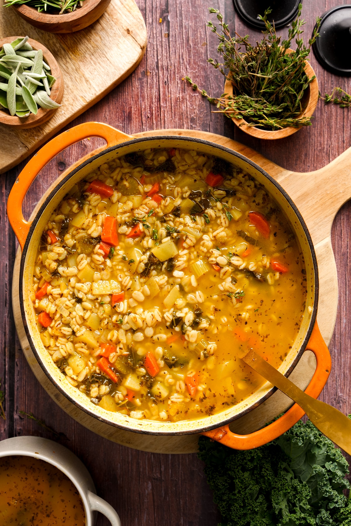 Overhead view of a Dutch oven filled with hearty Italian vegan barley soup made with carrots, potatoes, celery, and herbs in a golden broth, surrounded by fresh sage, thyme, and kale on a wooden table.
