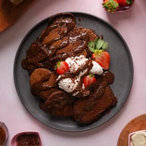 Vegan chocolate brownie pancakes topped with coconut whipped cream, chocolate drizzle, and fresh strawberries on a plate, an easy dairy-free plant-based breakfast recipe.
