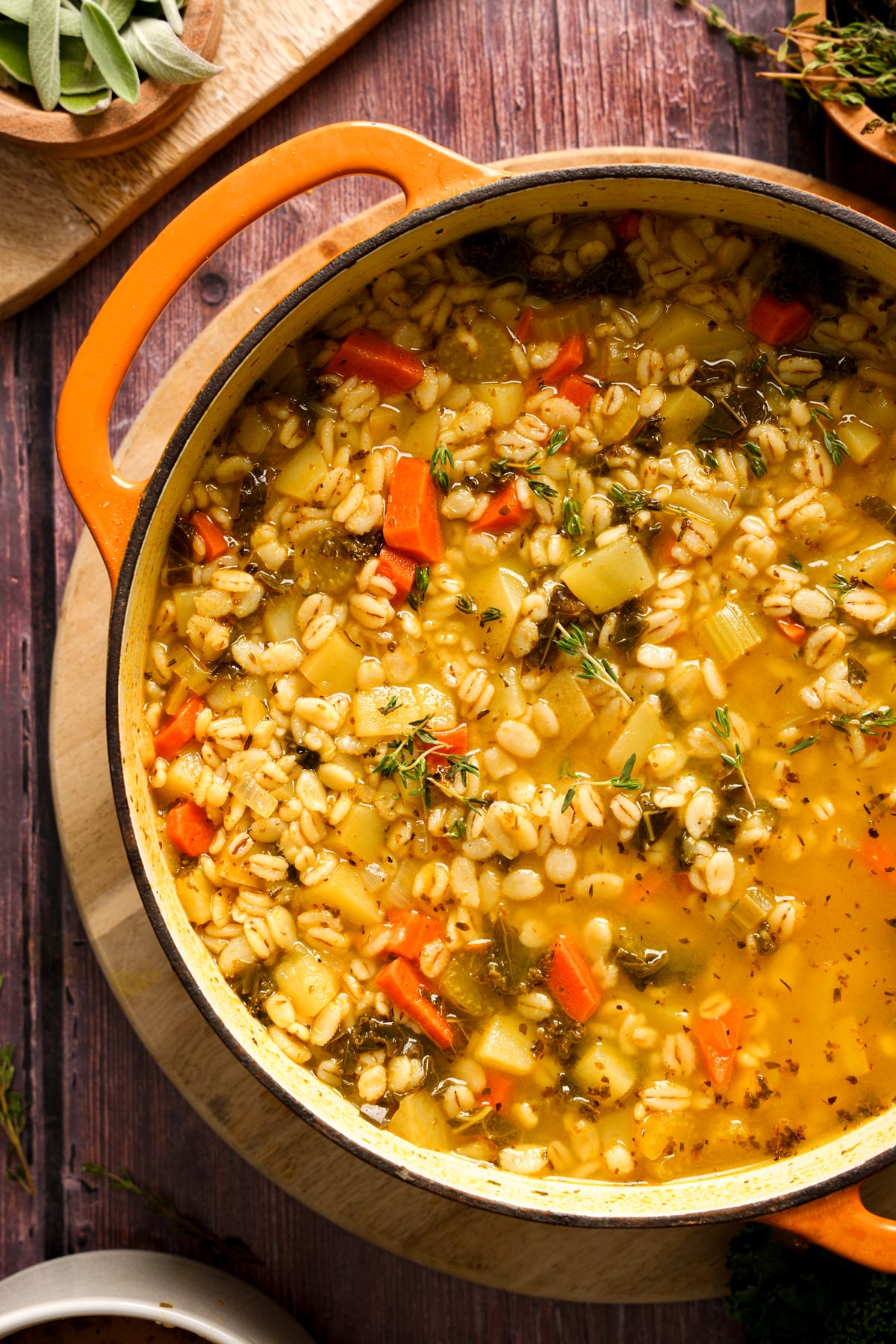 Large orange Dutch oven filled with hearty vegan barley soup made with carrots, potatoes, celery, and herbs simmering in a golden broth, served on a wooden board for a cozy plant-based meal.