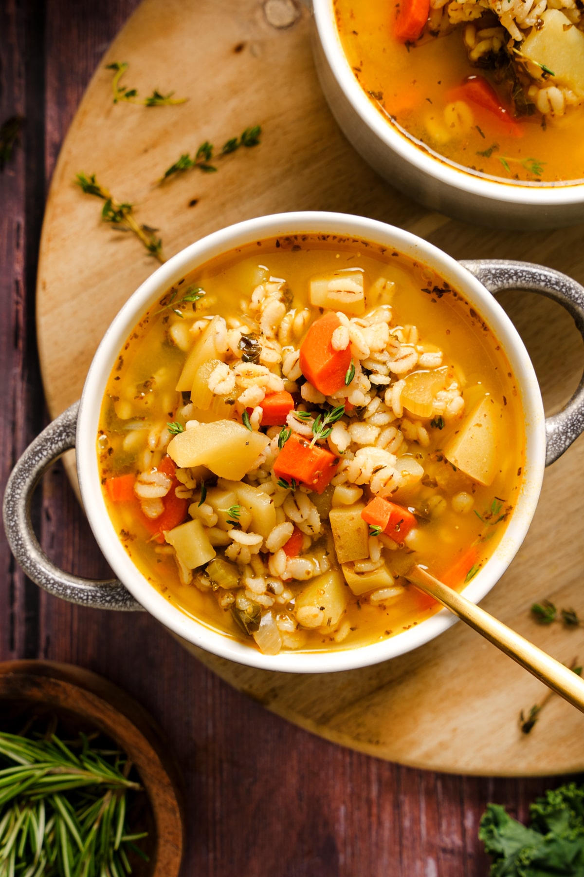 Bowl of Italian vegan barley soup filled with carrots, potatoes, celery, and fresh herbs in a golden broth, served with a gold spoon on a wooden board for a cozy plant-based meal.