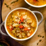 Bowl of hearty vegan barley soup filled with carrots, potatoes, celery, and fresh herbs in a golden vegetable broth, served on a wooden board for a cozy, comforting meal.