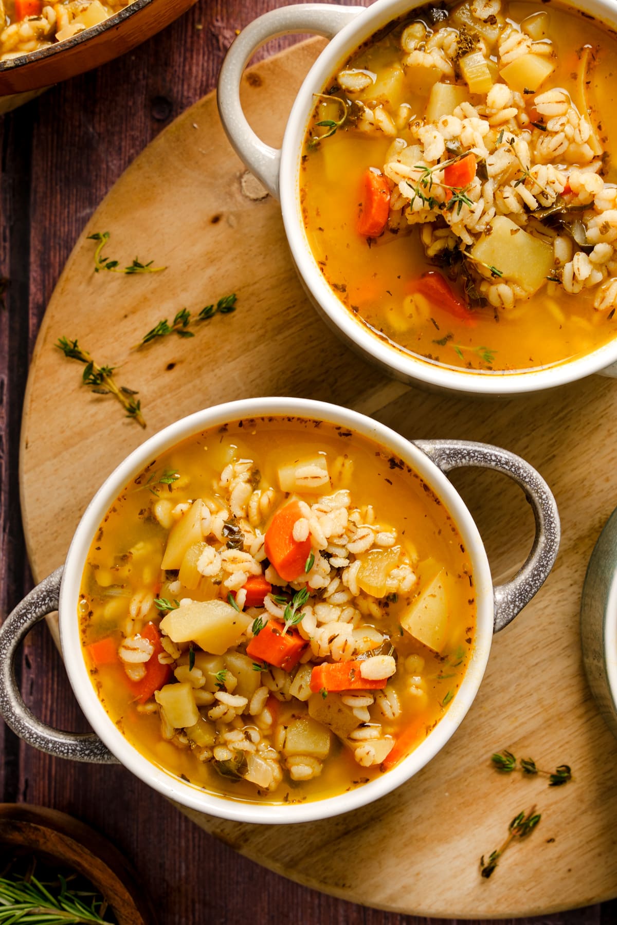 Two bowls of Italian vegan barley soup filled with carrots, potatoes, and fresh herbs in a golden broth, served on a wooden board for a cozy, plant-based meal.