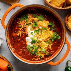 Top-down view of a hearty vegan bean chili in an orange Dutch oven, topped with vegan sour cream, shredded vegan cheese, tortilla chips, green onions, and fresh cilantro on a gray surface.