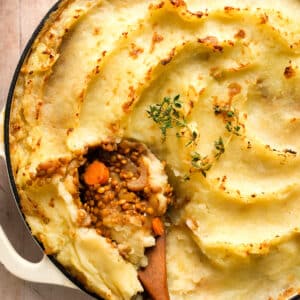 Close-up of vegan lentil shepherd’s pie with creamy mashed potato topping, a spoonful scooped out to reveal a hearty lentil and vegetable filling, garnished with fresh thyme in a white baking dish.