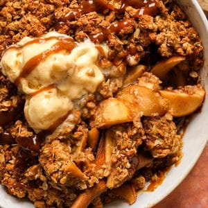 Close-up of healthy vegan apple crisp with tender cinnamon apples and a golden oat crumble, topped with melting vegan vanilla ice cream and caramel sauce.
