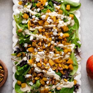 Close-up overhead view of a roasted golden beet salad on a white scalloped serving platter. The salad is made with mixed spring greens and topped with roasted golden beets, crunchy pistachios, crumbled vegan cheese, and a creamy dressing drizzled throughout. The colorful combination of greens, golden beets, and pistachios creates an elegant plant-based salad that's perfect for lunch, dinner, holidays, or entertaining.