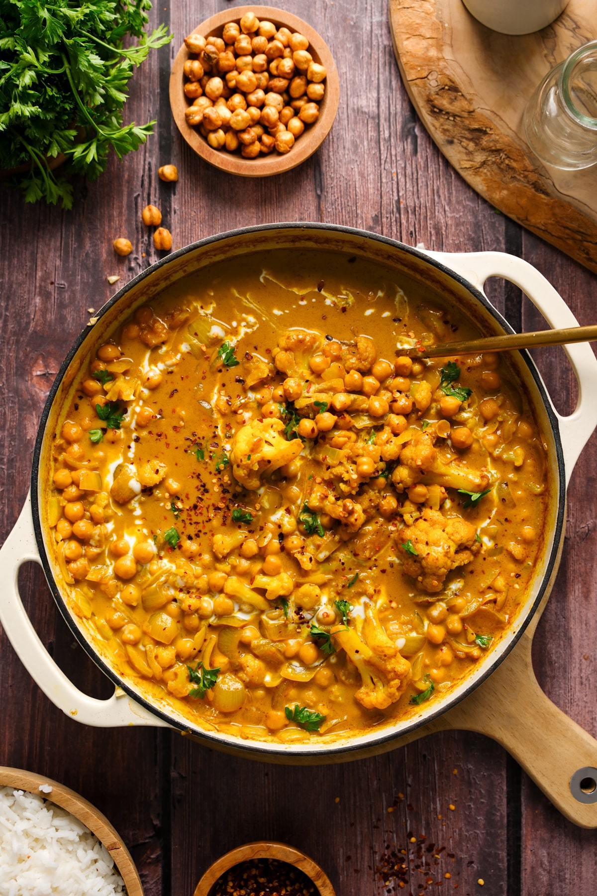 Overhead shot of a creamy vegan coconut chickpea curry simmering in a large cream-colored Dutch oven, filled with chickpeas, cauliflower, onions, and fresh herbs. The golden curry sauce is rich and glossy, swirled with coconut milk and garnished with red pepper flakes and parsley. Wooden bowls of rice, chili flakes, and crispy chickpeas are styled around the pot on a rustic dark wood table, enhancing the cozy, comforting presentation.