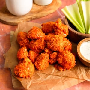 Overhead view of crispy vegan buffalo cauliflower bites coated in a bright, spicy buffalo sauce. The breaded cauliflower florets are piled high on parchment paper atop a wooden board and served with fresh celery sticks and a creamy dipping sauce. The vibrant orange coating and crunchy texture make this plant-based appetizer perfect for game day snacks, party food, or a healthy vegan comfort food recipe.