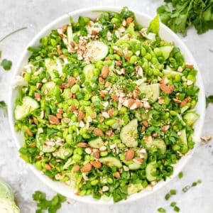 Overhead view of a large white bowl filled with a vibrant vegan edamame crunch salad, featuring chopped romaine, shredded cabbage, sliced cucumber, fresh cilantro, green onions, and crushed almonds, arranged on a light marble surface with scattered herbs and ingredients around the bowl.