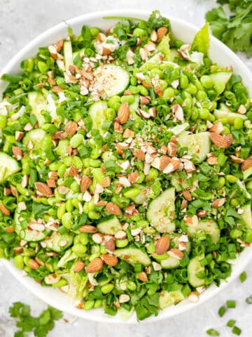 Overhead view of a large white bowl filled with a vibrant vegan edamame crunch salad, featuring chopped romaine, shredded cabbage, sliced cucumber, fresh cilantro, green onions, and crushed almonds, arranged on a light marble surface with scattered herbs and ingredients around the bowl.
