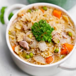 A close-up of a white bowl filled with vegan “chicken” noodle soup made with farfalle pasta, jackfruit, carrots, peas, celery, and onions, topped with a fresh parsley garnish.
