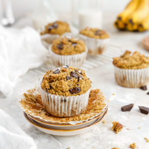 vegan chocolate chip banana muffins lined up and on a serving dish with crumbs and chocolate around them