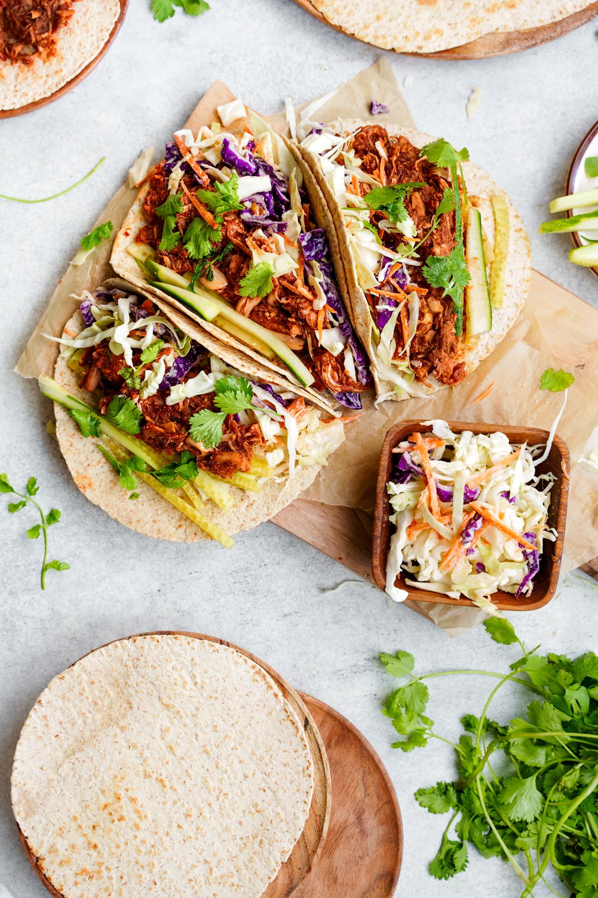 three bbq jackfruit tacos with a bowl of coleslaw on the side and plates with tortillas