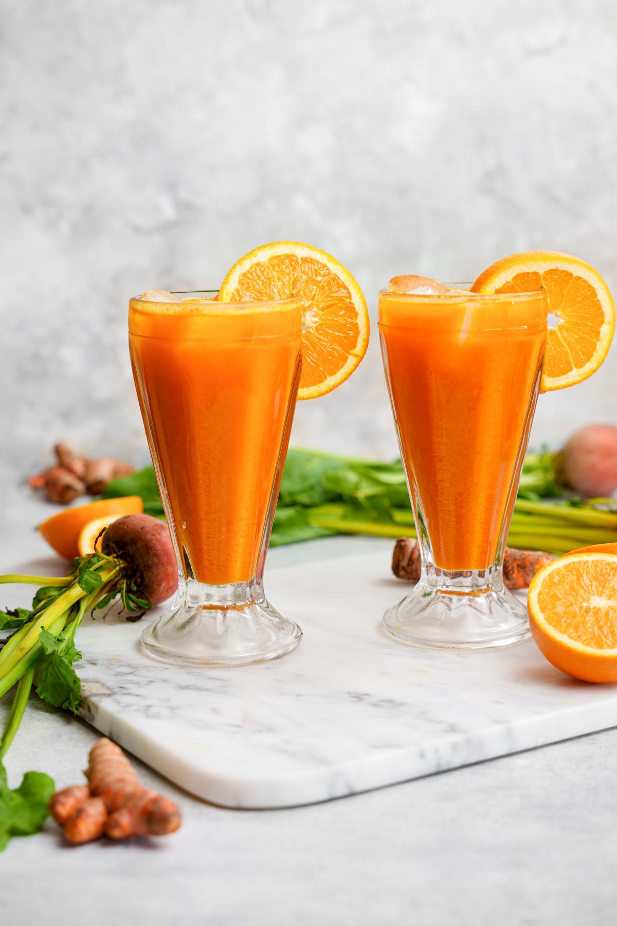 two glasses of the golden beet juice with fresh oranges, beets, and turmeric surrounding them