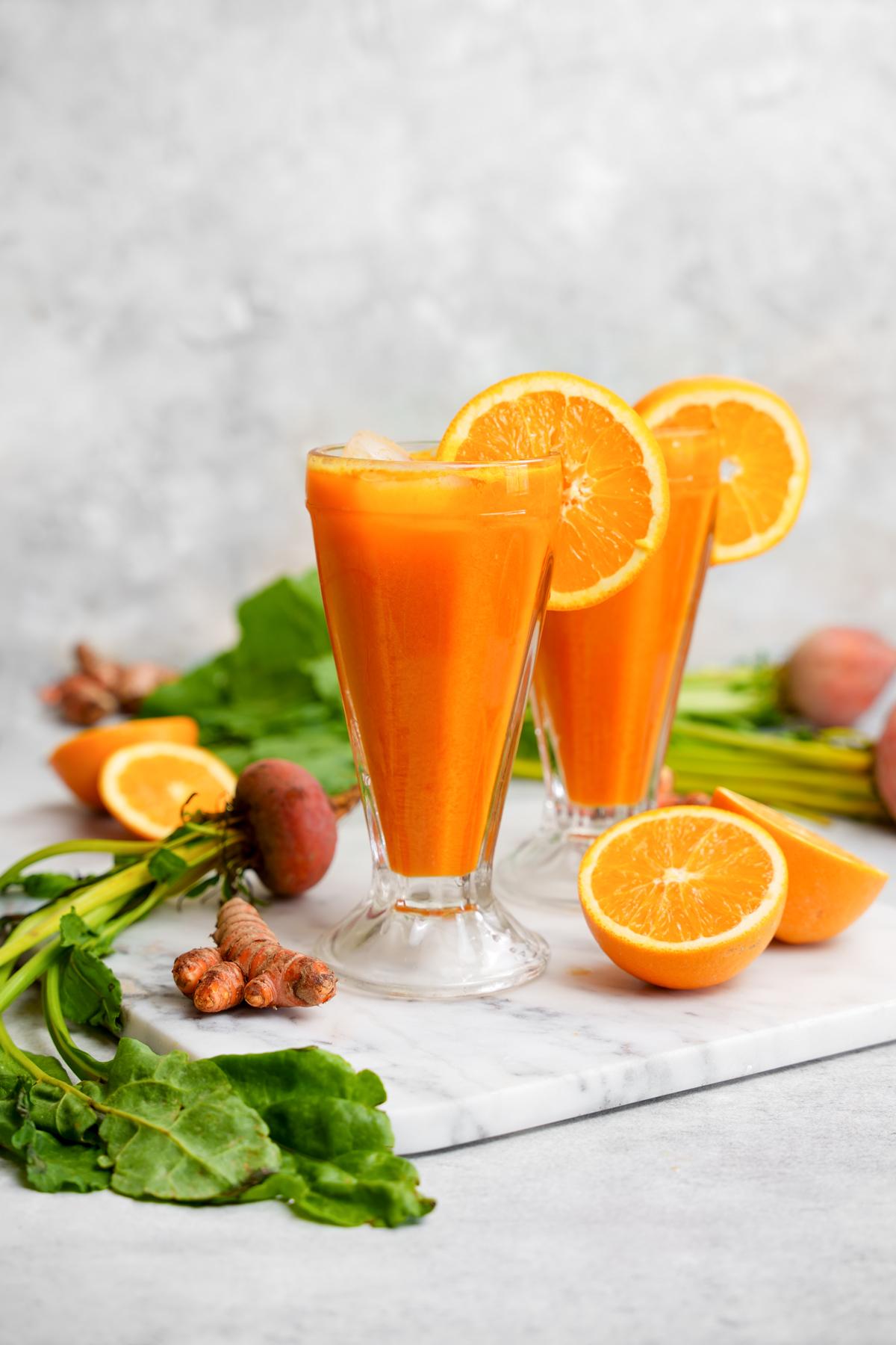 the golden beet juice in two glass cups surrounded by oranges, golden beets, and turmeric