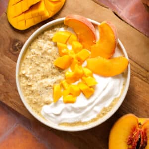 A bowl of healthy overnight mango peach oatmeal topped with creamy dairy-free yogurt, fresh diced mango, and juicy peach slices. Fresh honey mango and ripe peaches surround the bowl on a rustic wooden board, making a bright, easy make-ahead breakfast.
