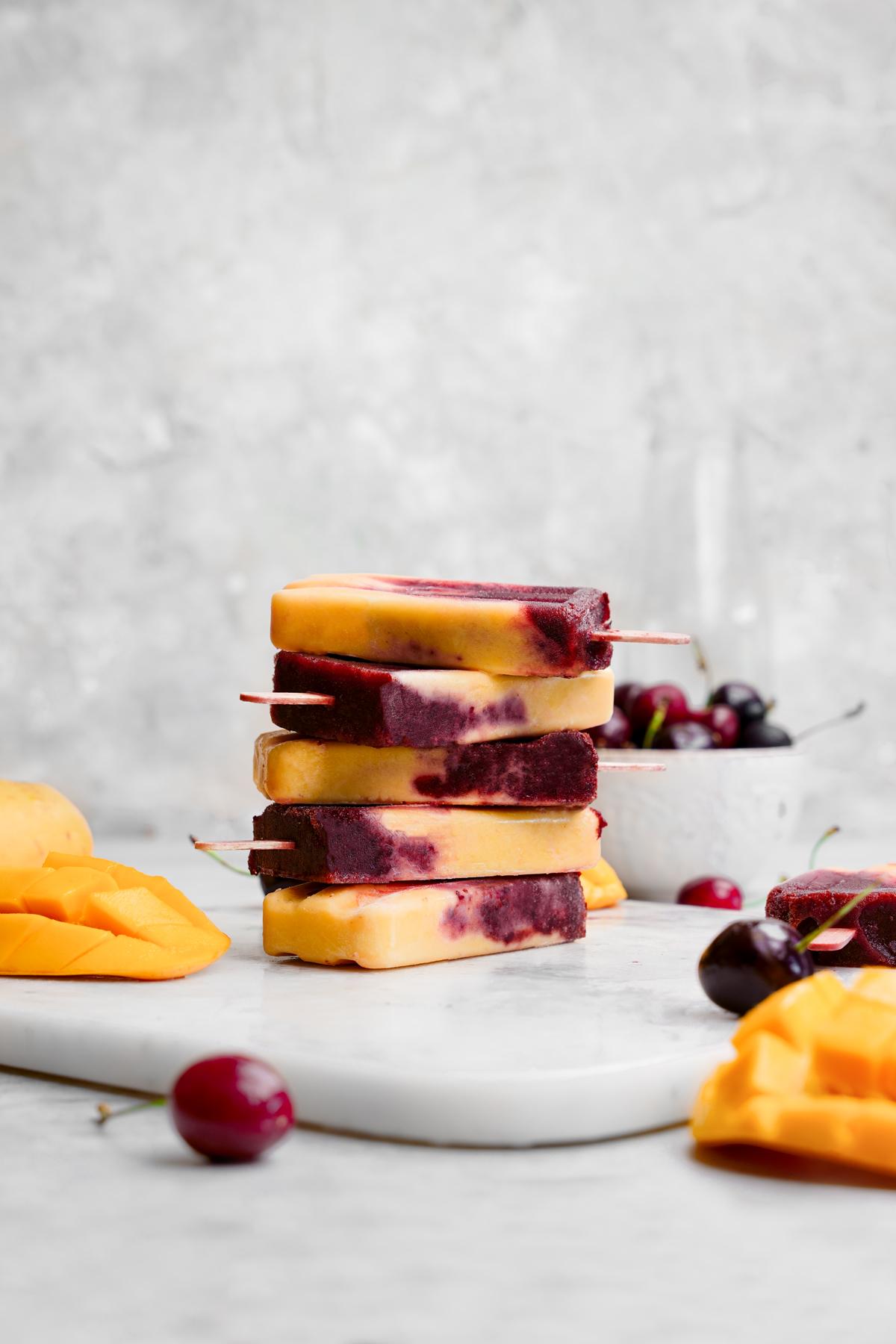 cherry mango popsicles stacked on top of each other with fresh mangos and cherries