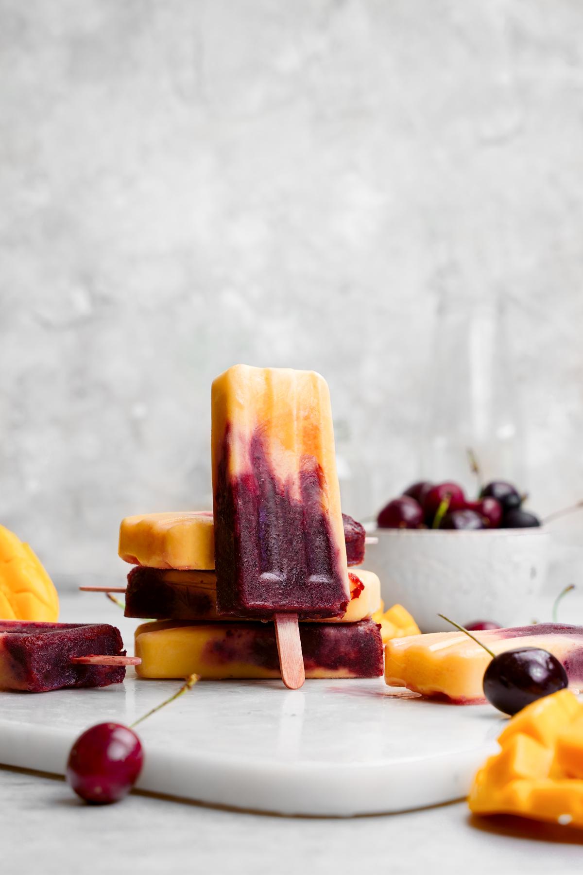 the cherry mango popsicles stacked on top of each other with one standing upright. The popsicles are surrounded by fresh ripe mango and cherries