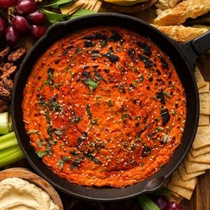 Overhead view of creamy vegan butternut squash dip baked in a cast iron skillet, topped with fresh sage, chives, sesame seeds, and seasoning, surrounded by crackers, bread, grapes, celery, and hummus.