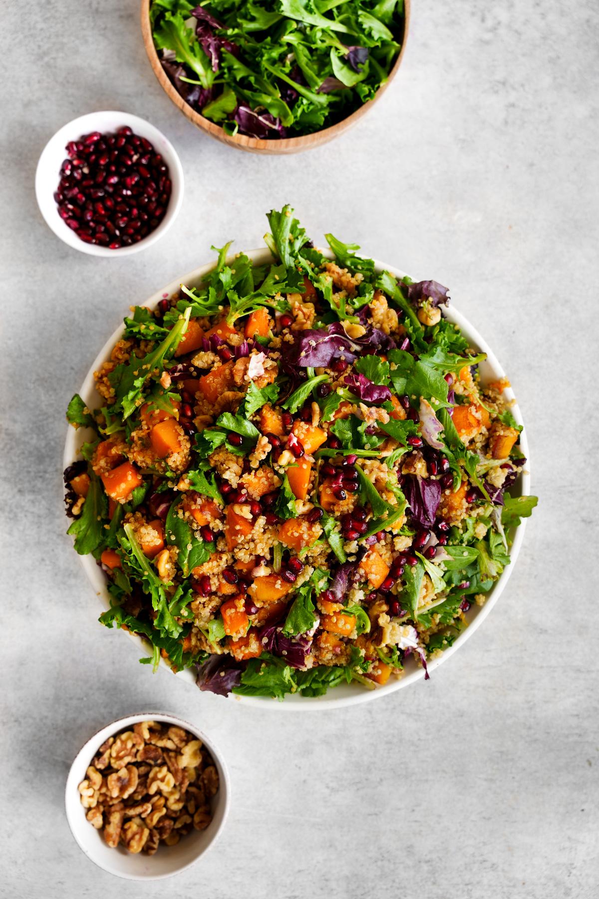 the butternut squash quinoa salad without any dressing and bowls of lettuce, walnuts, and pomegranates around it