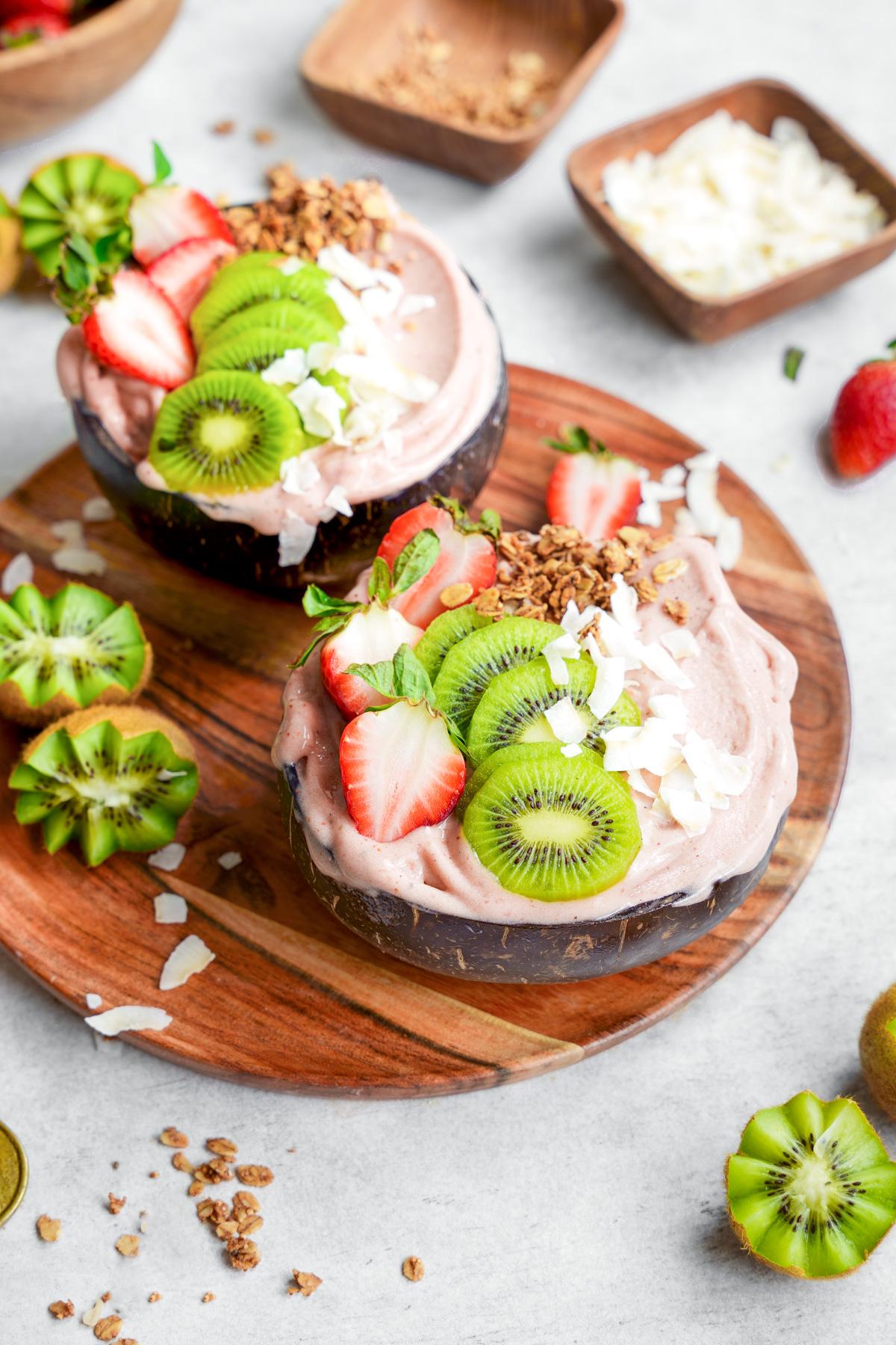 the strawberry kiwi smoothie bowls in coconut bowls with fresh ingredients on top and all around