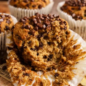 Close-up of a banana chocolate chip protein muffin broken open to reveal its soft, moist, fluffy interior packed with melted chocolate chips. The bakery-style muffin is topped with extra dairy-free chocolate chips and surrounded by crumbs, showcasing its tender texture. Made with ripe bananas and plant-based protein powder, these healthy vegan protein muffins are perfect for breakfast, meal prep, post-workout snacks, or a high-protein treat.