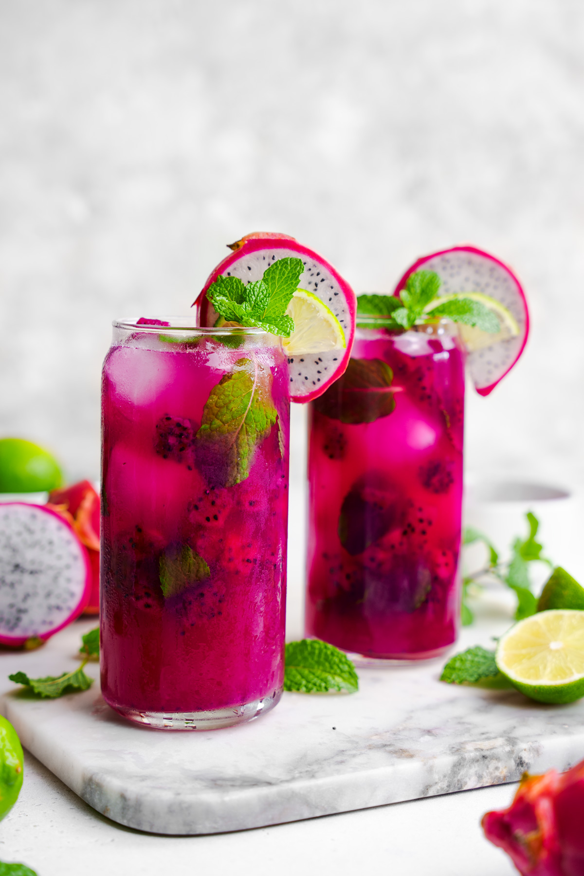 two glasses of the sol-ti dragon fruit mojito mocktails together with fresh limes, mint, and dragon fruit surrounding them.