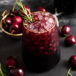 Close-up of a ruby red Sleepy Girl Mocktail filled with crushed ice and garnished with fresh rosemary, surrounded by fresh cherries.