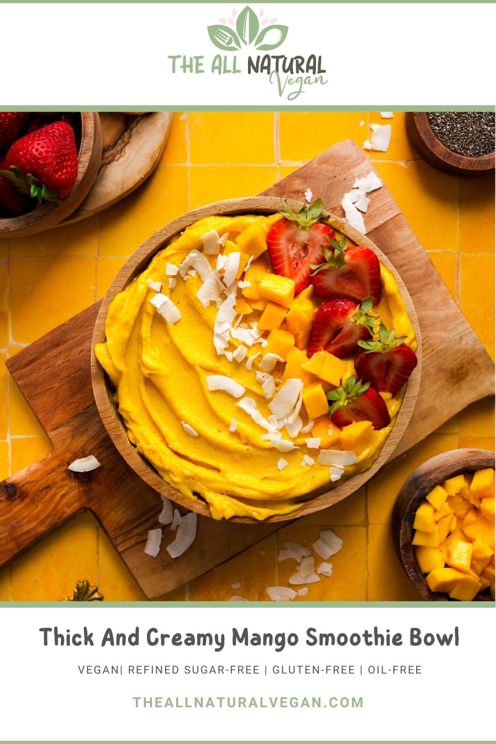 Thick and creamy mango smoothie bowl topped with diced mango, fresh strawberries, and coconut flakes, served in a wooden bowl on a cutting board over a bright yellow tile background. Labeled as vegan, gluten-free, refined sugar-free, and oil-free from The All Natural Vegan.