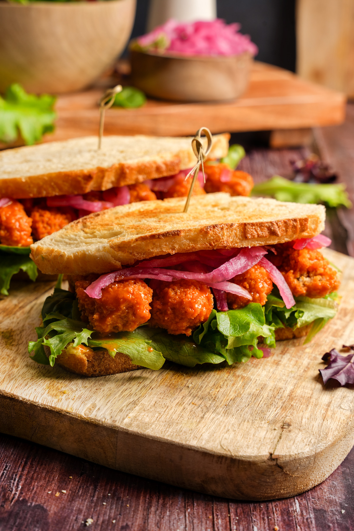 Two vegan buffalo tempeh sandwiches on toasted bread, layered with leafy green lettuce, tangy pickled red onions, and saucy buffalo tempeh bites, served on a rustic wooden cutting board.