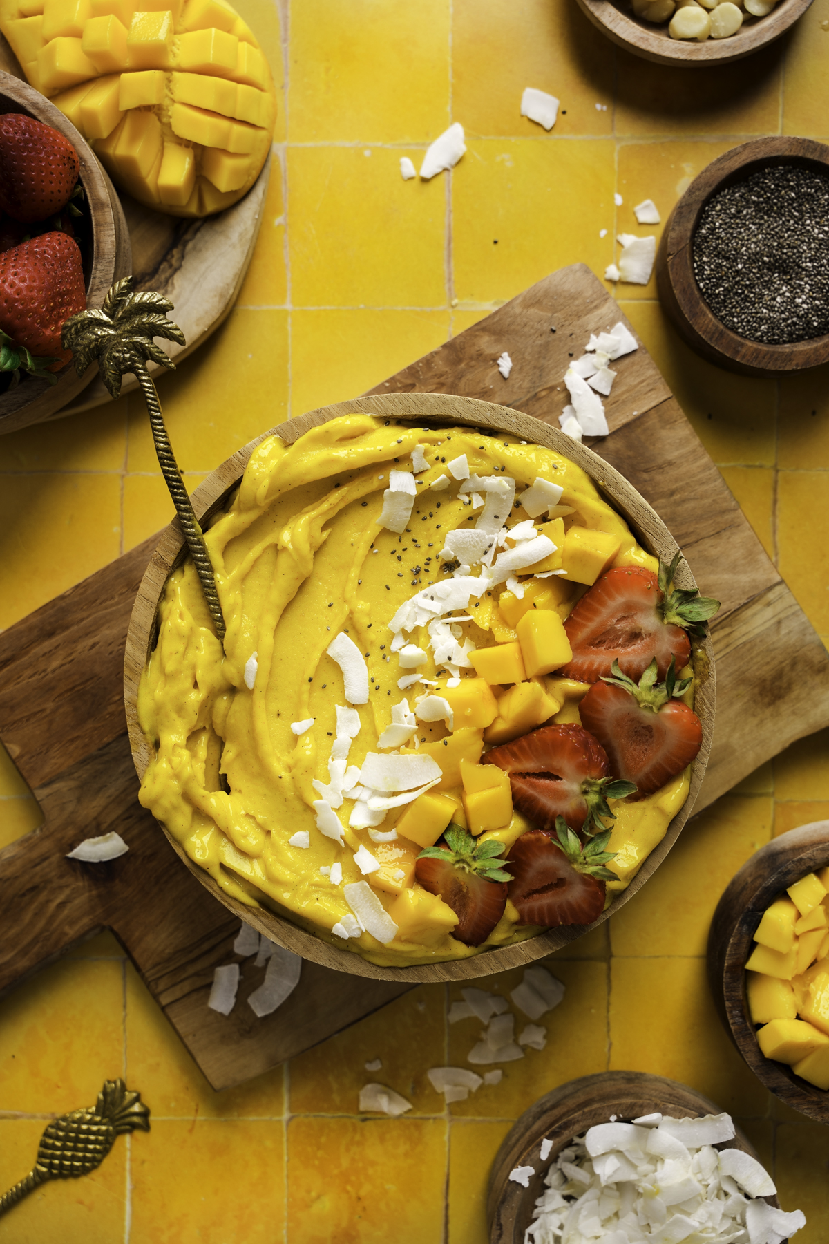 Vibrant mango smoothie bowl topped with diced mango, sliced strawberries, chia seeds, and coconut flakes, served in a wooden bowl with a gold spoon on a bright yellow tile background surrounded by fresh fruit and toppings.