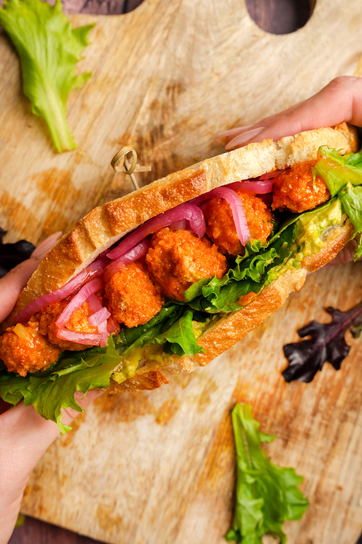 Close-up of a hand holding a toasted vegan buffalo tempeh sandwich filled with fresh leafy greens, smashed avocado, tangy pickled red onions, and crispy buffalo tempeh bites on rustic bread.