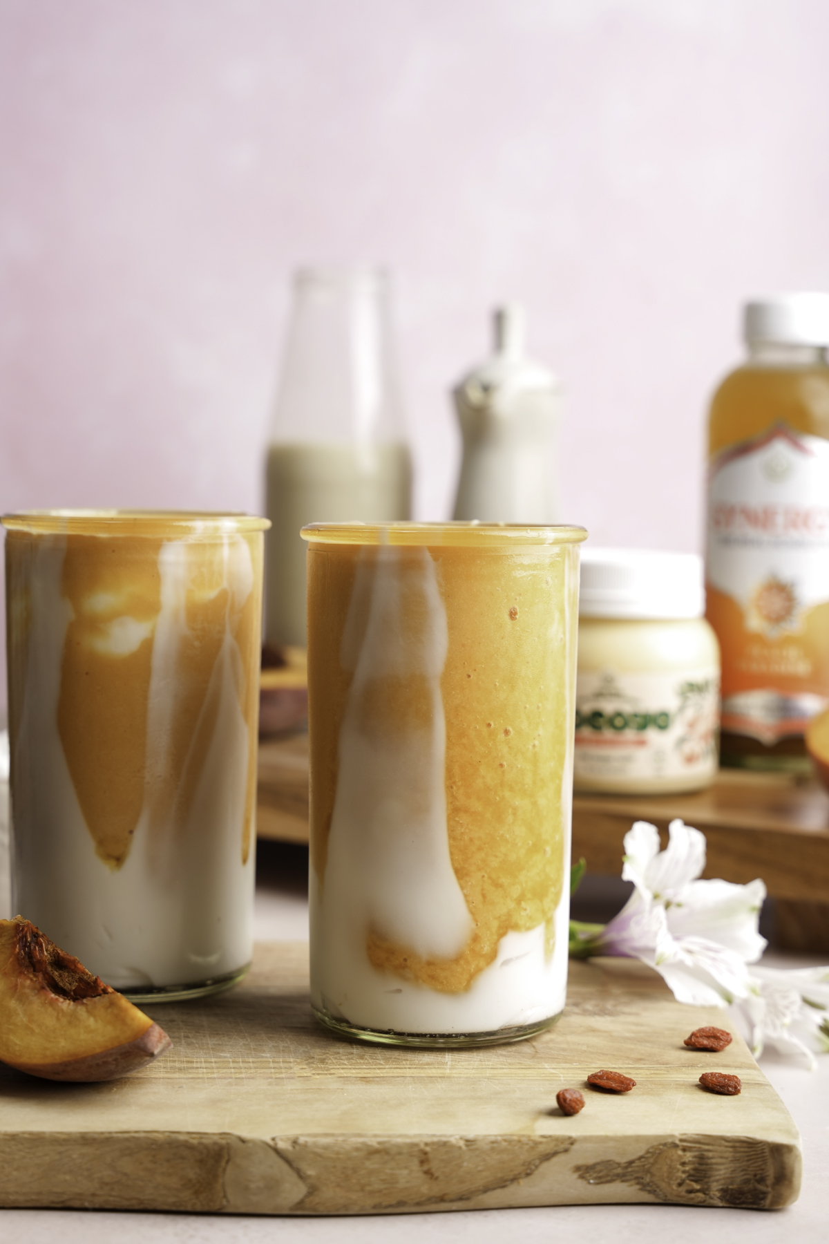 Peaches and cream smoothie made with coconut yogurt and fresh peach puree, served in tall clear glasses with a swirl effect. Set on a wooden board with peach slices, goji berries, and probiotic-rich ingredients in the background.