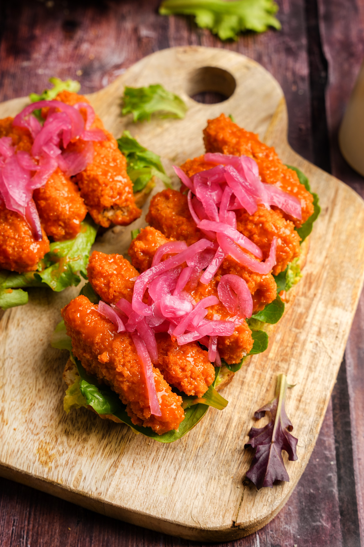 Open-faced vegan buffalo tempeh sandwiches on a wooden cutting board, topped with crispy lettuce, spicy buffalo-coated tempeh strips, and vibrant pickled red onions.
