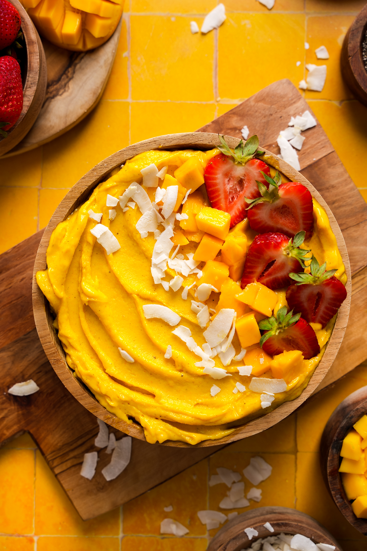 Vegan mango smoothie bowl topped with fresh diced mango, halved strawberries, and coconut flakes, served in a wooden bowl on a yellow tiled surface with scattered toppings.