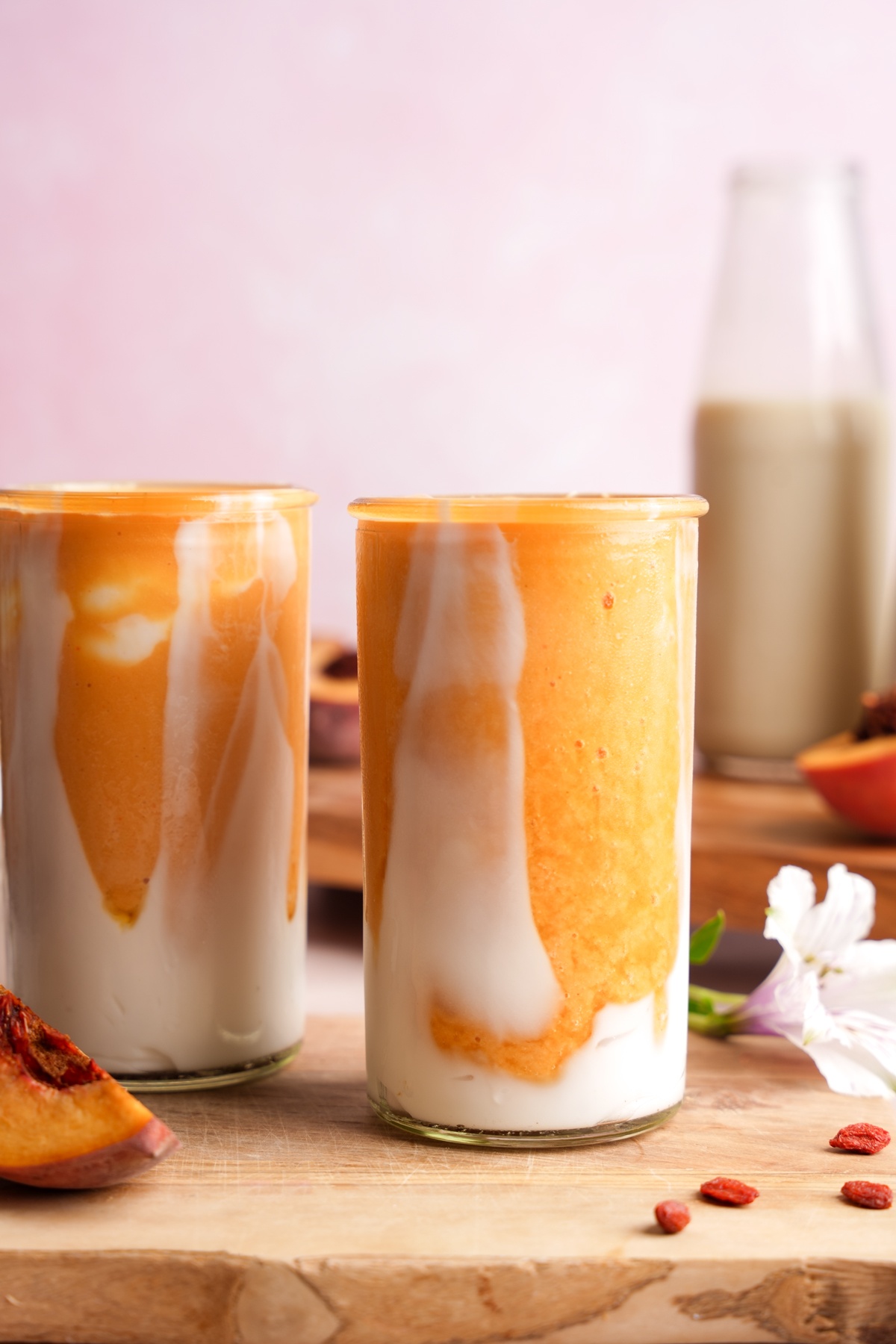 Vegan peaches and cream smoothie in tall glasses, featuring a swirl of creamy coconut yogurt and peach smoothie, set on a wooden board with fresh peach slices, goji berries, and a pink background.