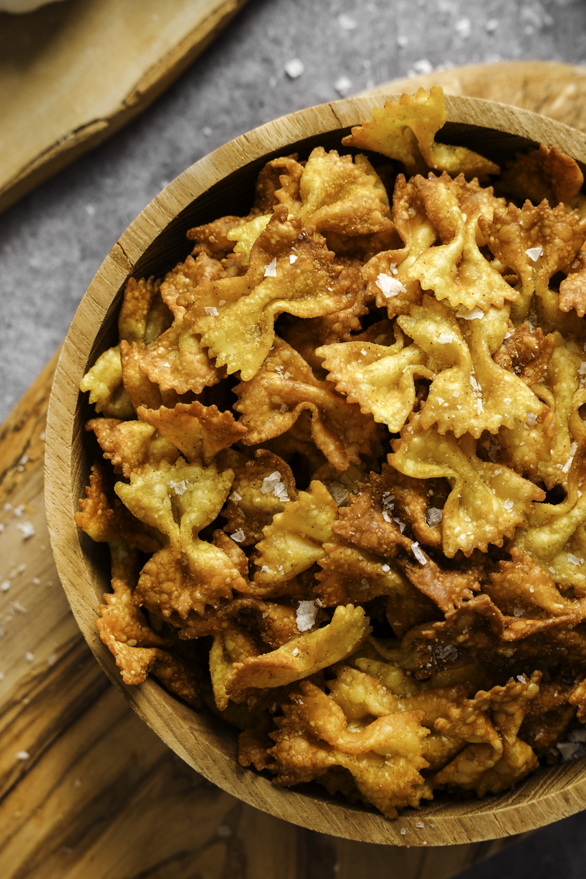 Close-up of a wooden bowl filled with golden, crispy bowtie pasta chips, lightly dusted with flaky sea salt. The pasta is evenly fried or baked to a crunchy texture, showcasing a mix of light and golden-brown tones, sitting on a wooden board over a gray stone surface with scattered salt crystals.