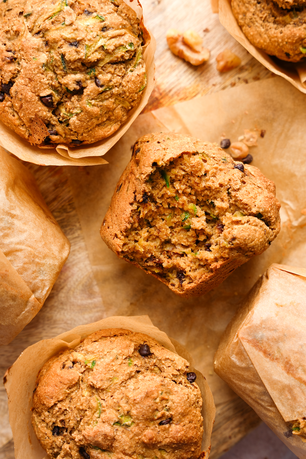 Moist vegan zucchini muffins with chocolate chips, shown on parchment paper with one muffin broken open to reveal a soft, tender crumb and bits of shredded zucchini.