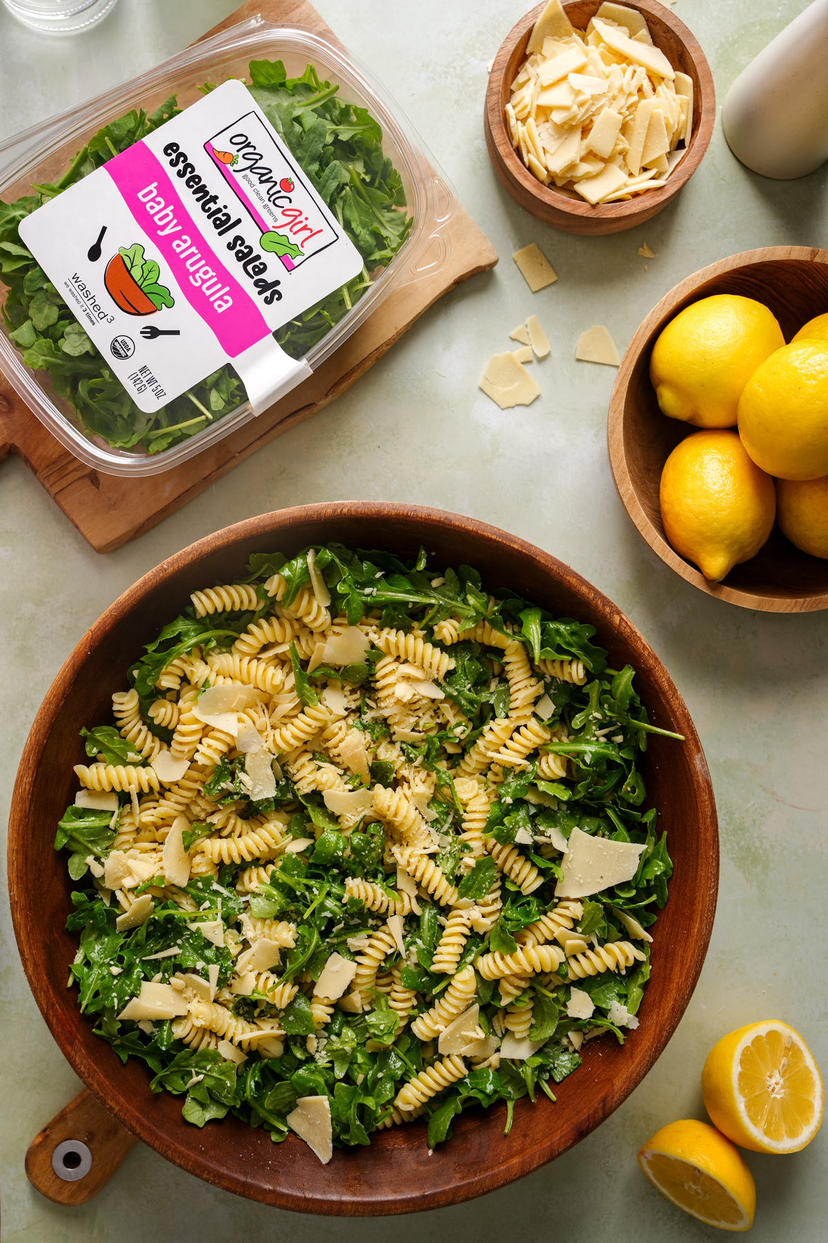 Overhead view of a wooden bowl filled with arugula pasta salad made with rotini pasta, organicgirl baby arugula, shaved vegan parmesan, and a lemon vinaigrette, surrounded by fresh lemons and arugula packaging on a light countertop.