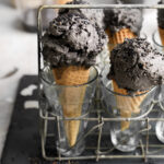 Close-up of two waffle cones filled with rich, dark black sesame ice cream, topped with black sesame seeds, displayed upright in clear glasses within a rustic wire rack.