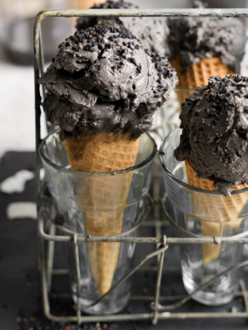 Close-up of two waffle cones filled with rich, dark black sesame ice cream, topped with black sesame seeds, displayed upright in clear glasses within a rustic wire rack.