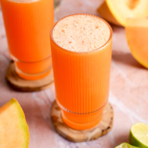 Vibrant glass of cold-pressed cantaloupe juice topped with natural foam, sitting on a wooden coaster, surrounded by fresh cantaloupe wedges and lime halves on a soft pink surface.