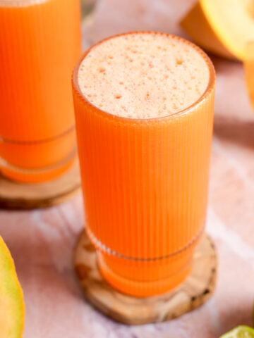 Vibrant glass of cold-pressed cantaloupe juice topped with natural foam, sitting on a wooden coaster, surrounded by fresh cantaloupe wedges and lime halves on a soft pink surface.