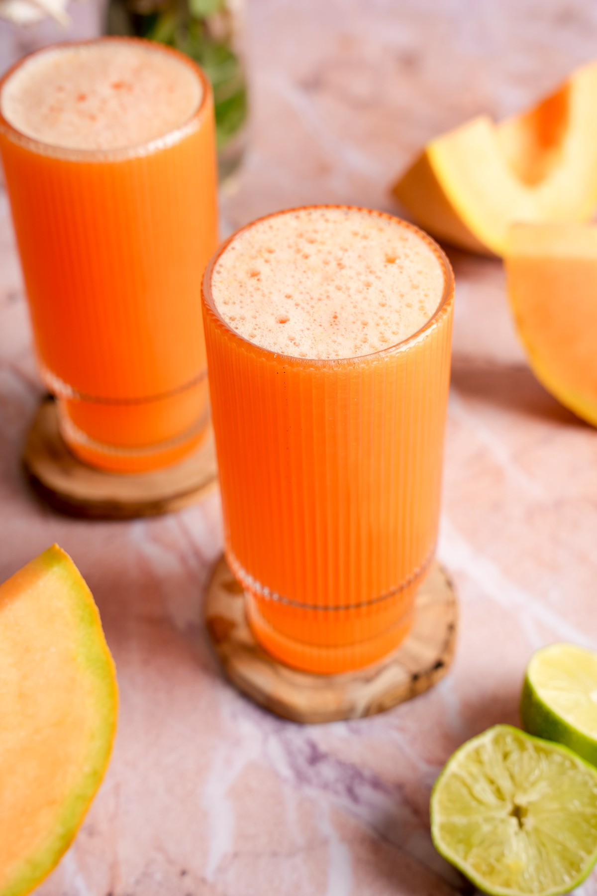 Freshly cold-pressed cantaloupe juice served in tall ridged glasses, surrounded by ripe cantaloupe wedges and halved limes on a pink marbled background.