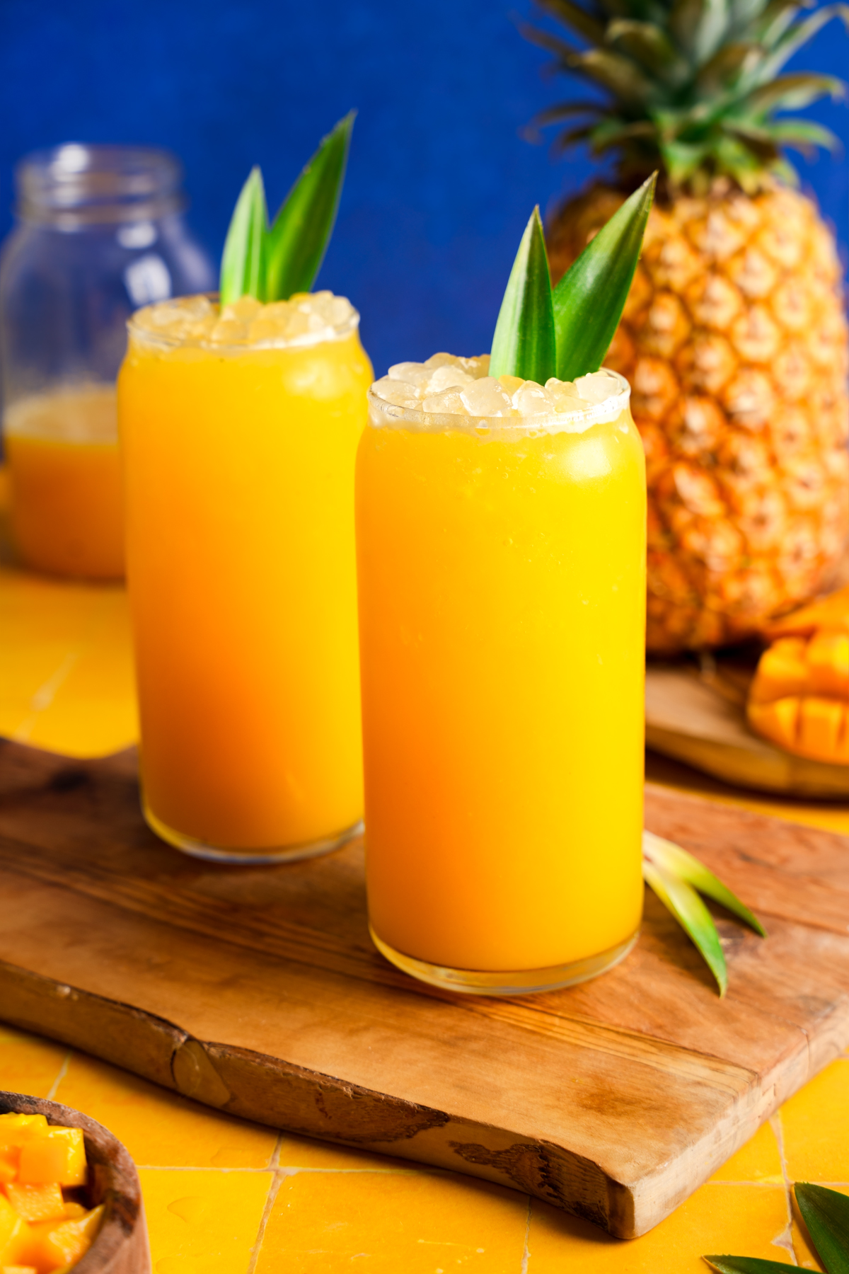 Two glasses of fresh mango pineapple juice topped with crushed ice and garnished with pineapple leaves, set on a wooden board with a ripe pineapple, mango chunks, and a blue background for a vibrant tropical presentation.