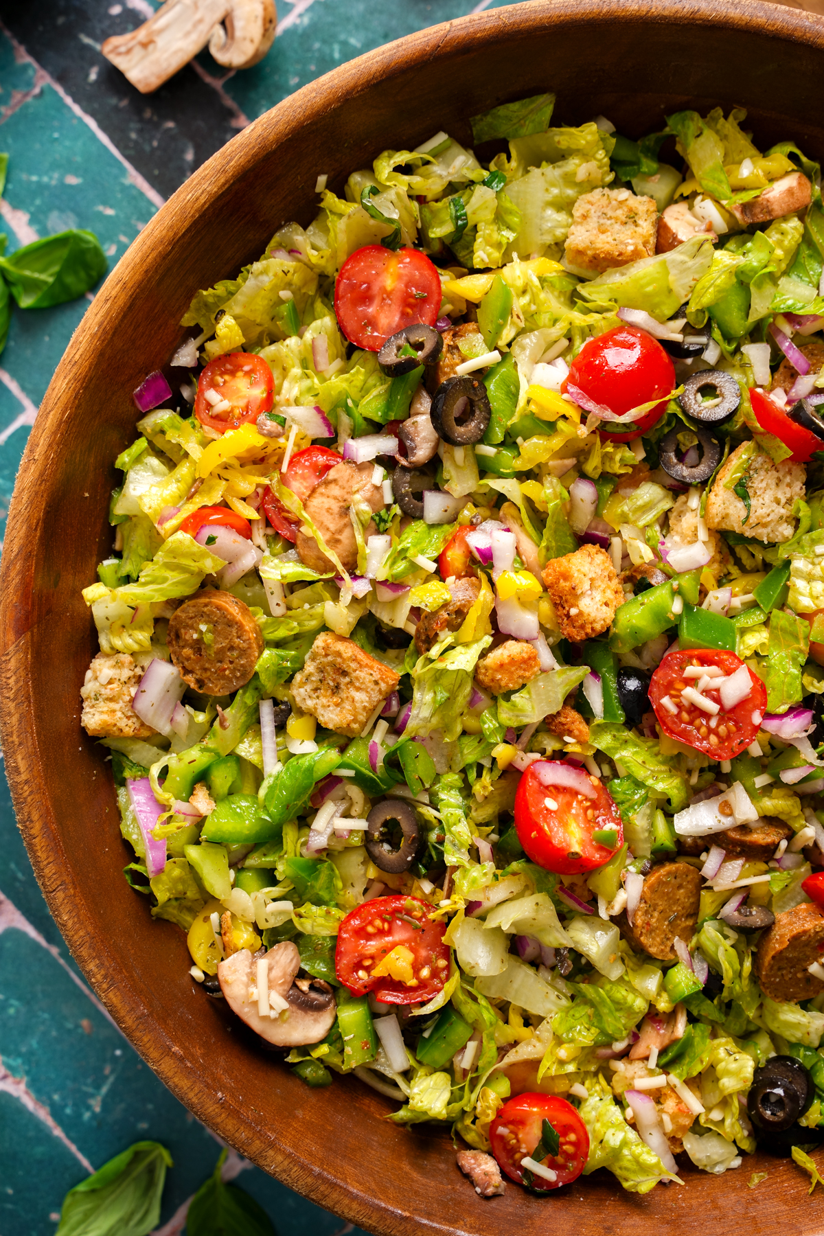 Close-up of a vibrant vegan pizza salad in a wooden bowl featuring chopped romaine, cherry tomatoes, black olives, banana peppers, vegan sausage, red onions, mushrooms, croutons, and shredded vegan cheese