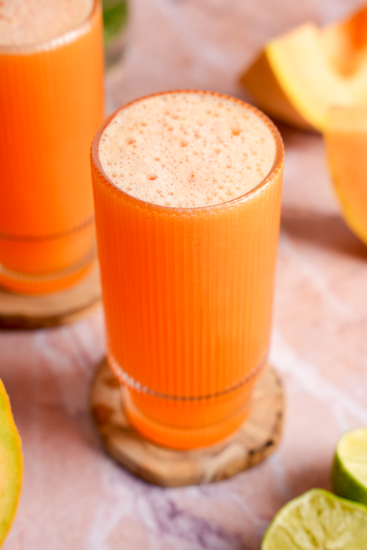 Freshly made cold-pressed cantaloupe juice in a tall, ribbed glass on a wooden coaster, surrounded by ripe cantaloupe slices and halved limes