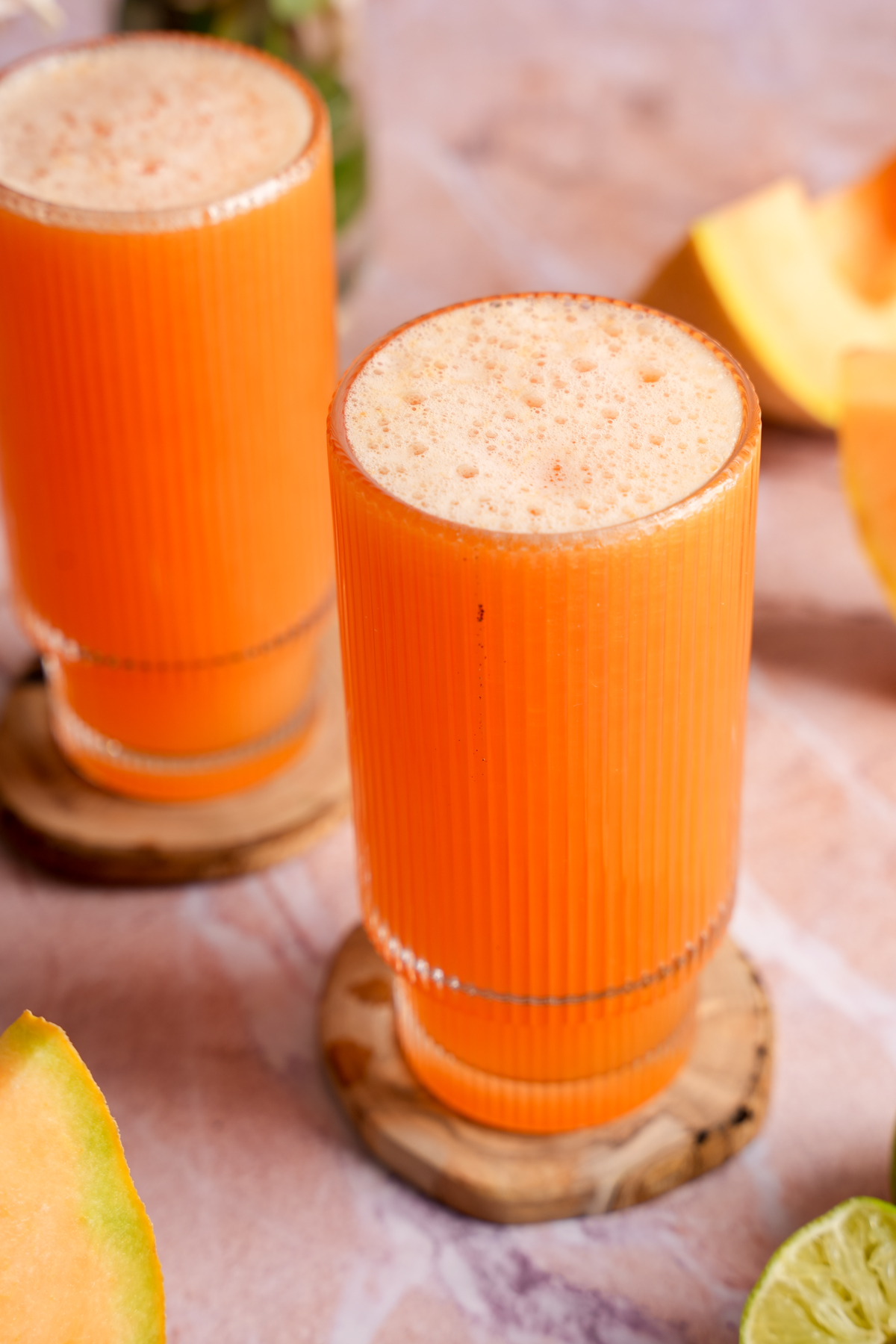 Tall glass of vibrant orange cantaloupe juice with a light foam layer on top, placed on a wooden coaster and surrounded by fresh lime halves and cantaloupe wedges on a pink marble surface.