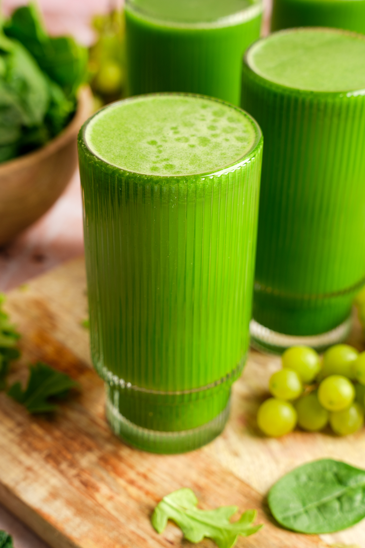 Close-up of a vibrant green juice served in a tall ridged glass, surrounded by fresh spinach, arugula, and green grapes on a wooden board — healthy detox green juice made with kale, cucumber, green apples, and leafy greens.